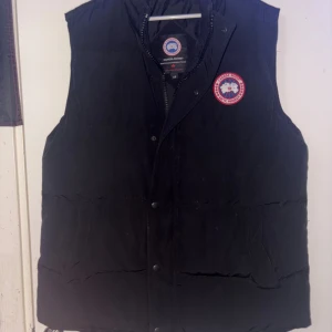 Svart dunväst från Canada Goose - Svart dunväst från Canada Goose med klassisk logga på bröstet. Västen har både dragkedja och tryckknappar framtill, samt en quiltad design som ger extra värme. Perfekt för kalla dagar och har en stilren look som funkar till många outfits.