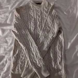 Beige stickad kabeltröja Lager 157 - Snygg beige stickad tröja från H&M med klassiskt kabelmönster. Tröjan har rund hals, långa ärmar och ribbade muddar vid ärmslut och nederkant. Perfekt för dig som gillar en chill och stilren look under höst och vinter.