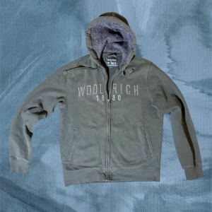 Mörkgrön hoodie från Woolrich - Mysig mörkgrön hoodie från Woolrich med broderad logga och årtal framtill. Tröjan har huva med snörning, mjukt foder och praktiska fickor. Perfekt för kyliga dagar och enkel att matcha med jeans eller joggers.