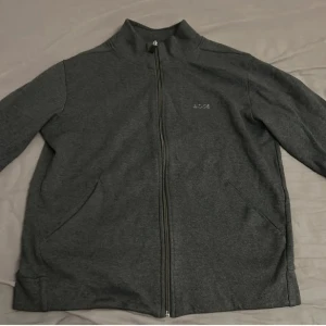 Mörkgrå zipjacka från BOSS - Snygg mörkgrå zipjacka från BOSS med stilren design och diskret logga på bröstet. Jackan har hel dragkedja, ståkrage och två sidofickor. Tillverkad i mjuk bomullsmix som är skön att bära. Perfekt för chill dagar eller när du vill ha en clean look.