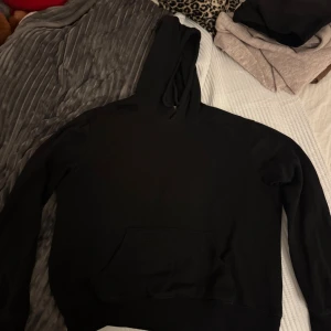 Svart hoodie med ficka, storlek S - Svart hoodie i klassisk stil med stor magficka och huva. Tröjan har långa ärmar och en relaxed passform som är perfekt för chill dagar. Materialet känns mjukt och skönt mot huden, och den är enkel att matcha med jeans eller joggers.