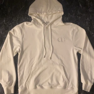 Vit CD hoodie med broderad logga - Säljer en stilren vit hoodie med broderad CD-logga på bröstet. Tröjan har huva med snörning och en stor magficka. Perfekt för dig som gillar exklusiva och minimalistiska plagg.