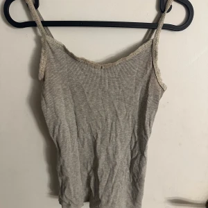 Grå ribbad linnetopp från H&M - Säljer en ljusgrå ribbad linnetopp från H&M i storlek 158/164. Toppen har tunna axelband och spetskant längs halsringningen för en söt touch. Materialet är mjukt och stretchigt, perfekt för en chill look. Passar dig som gillar enkla och stilrena plagg.