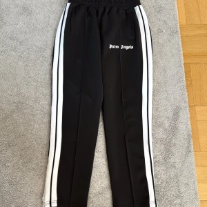 Palm Angels svarta mjukisbyxor  äkta - Svarta mjukisbyxor från Palm Angels med vita dubbla sidoremsor och logotyp broderad på framsidan. Byxorna har elastisk midja med snörning och raka ben. Materialet är mjukt och stretchigt, perfekt för chill eller streetwear. Snygga fickor på sidorna.