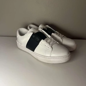 Valentino Garavani Open sneakers vit/svart - Snygga Valentino Garavani Open sneakers i vitt skinn med bred svart läderdetalj över vristen. Klassisk rund tå, vita snören och ikoniska nitar längs hälen. Perfekta för dig som gillar stilrena och lyxiga sneakers med en twist.