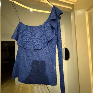 Blå spetsklänning med volang och oneshoulder💙 - Snygg blå kortklänning i spets med blommigt mönster. Klänningen har en axelbandslös design med en lång ärm och volangdetalj över bysten. Perfekt för dig som vill sticka ut med en unik och trendig look.💙