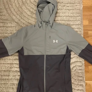 Under armour jacka - Snygg tvåfärgad vindjacka från Under Armour i grått och mörkgrått. Jackan har huva, hel dragkedja framtill, en bröstficka med dragkedja och elastiska muddar med logotypdetalj. Perfekt för träning och chill utomhus.