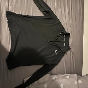 Svart Nike Dri-Fit half zip tröja - Svart långärmad tröja från Nike med Dri-Fit material, perfekt för löpning och träning. Tröjan har en vit Nike-logga på bröstet och en praktisk half zip framtill. Snygg och sportig passform som sitter bekvämt under aktivitet.