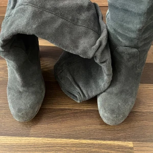 Grå mockastövlar med klack - Snygga grå stövlar i mocka med hög klack och rund tå. Stövlarna har en slouchy passform och träfärgad klack som ger en cool och trendig look. Perfekta att styla med jeans eller kjol för en extra touch till outfiten.