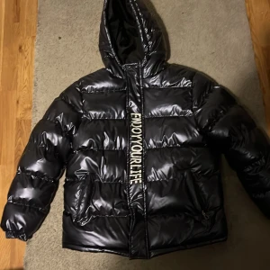 Svart pufferjacka med tryck - Svart, glansig pufferjacka med huva från Fashion Kids CYFS. Jackan har vita tryck på både fram- och baksidan, bland annat texten 'ENJOY YOUR LIFE' längs dragkedjan. Utrustad med två fickor och långa ärmar. Perfekt för dig som gillar enkel o clean style