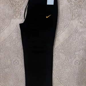 Nike NOCTA svarta mjukisbyxor - Helt nya svarta mjukisbyxor från Nike NOCTA i storlek S med gul logga och detaljer. Byxorna har dragsko i midjan. Tillverkade i mjukt material för maximal komfort och stilren look. Byxorna är oanvända och taggarna sitter kvar.