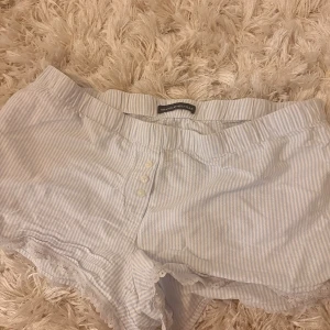 Randiga shorts från Brandy Melville - Söta ljusblå och vita randiga shorts från Brandy Melville med knappar framtill och spetsdetaljer längs bensluten. One size. Så sköna