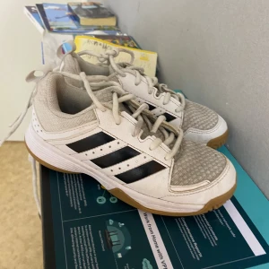 Vita Adidas sneakers med svarta ränder - Säljer ett par fräscha Adidas sneakers i vitt med klassiska svarta ränder och ljusbrun sula. Skorna har snörning och meshdetaljer framtill för extra andningsförmåga. Perfekta för dig som gillar sportig stil och vill ha bekväma skor till vardagen.