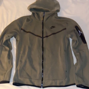 Olivgrön nike tech fleece Small - Olivgrön fleecejacka från Nike med huva och hel dragkedja framtill. Jackan har svarta detaljer, inklusive Nike-logga på bröstet och en ficka med dragkedja på ärmen. Perfekt för chill dagar och sportiga outfits. Katthår kan förekomma.