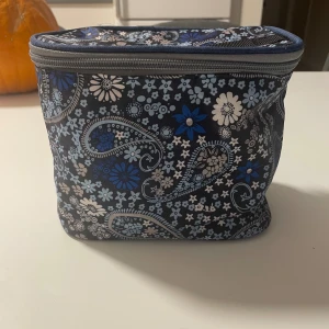 Blommig blå nessecär med dragkedja - Säljer en blommig nessecär i blå och grå toner med paisley-mönster. Väskan har en fyrkantig form och stängs med dragkedja upptill. Materialet är mjukt syntet och insidan är ljusgrå. Perfekt för att hålla ordning på småsaker.