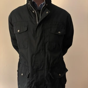 Field jacka  - Svart field jacka med fyra stora fickor framtill och snygga metallknappar. Jackan har hög krage, dragkedja och tryckknappar. Patch med logga på ena ärmen. Perfekt för dig som gillar stilren och funktionell design. Sitter som m fast är L