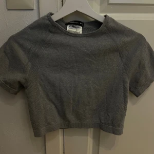 Grå croppad träningstopp från New Yorker - Säljer en grå croppad träningstopp från New Yorker med korta ärmar och rund hals. Toppen har en tight passform och är tillverkad i ett mjukt syntetmaterial som andas, perfekt för gymmet eller sport. Enkel och stilren design som funkar till alla träningsoutfits.