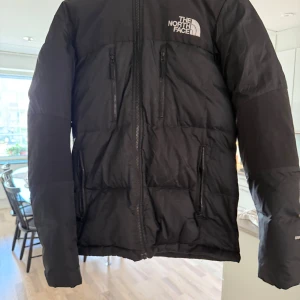 Svart pufferjacka från The North Face - Säljer en svart pufferjacka från The North Face med huva och flera dragkedjeförsedda fickor. Jackan har en quiltad design och logga på både bröstet och ryggen. Perfekt för kalla dagar och riktigt snygg streetstil.