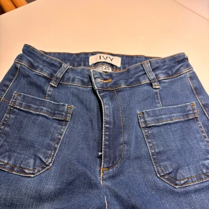 Blå bootcut jeans från IVY, stl 25 - Snygga blå jeans från IVY med bootcut-ben och hög midja. Klassisk femficksmodell med markerade sömmar och bälteshällor. Jeansen är tillverkade i jeansmaterial och har en tidlös blå tvätt som passar till allt.