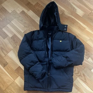 Svart pufferjacka från Lyle & Scott - Svart pufferjacka från Lyle & Scott med huva och gul logga på bröstet. Jackan har quiltad design, dragkedja framtill och långa ärmar. Perfekt för kalla dagar och har en clean, stilren look som funkar till det mesta.