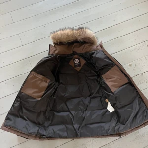 Brun Canada Goose dunjacka - Säljer en brun dunjacka från Canada Goose med fet pälskant på huvan och klassisk logga på ärmen. Jackan har två stora fickor framtill, dragkedja och quiltad design. Perfekt för kalla dagar och har en riktigt schysst look.