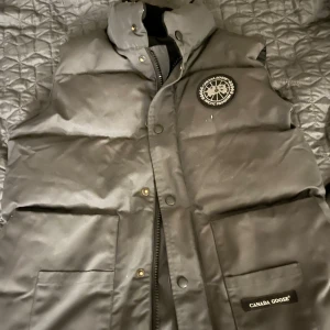 Svart dunväst från Canada Goose - Svart dunväst från Canada Goose med hög krage, tryckknappar och dragkedja framtill. Västen har två stora fickor med lock och en rund logotyp på bröstet. Perfekt för kalla dagar och har en stilren, klassisk look.