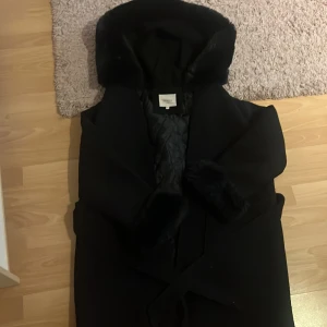 Svart kappa med fuskpäls från Orcelly - Svart kappa från Orcelly med bred krage och fuskpälsdetaljer på krage och ärmslut. Jackan har quiltat innerfoder, bälte i midjan och en stilren, lång siluett. Perfekt för dig som vill ha en snygg och varm look under höst och vinter.jag är storlek xs/s men köpte den två storlekar större och den satt bra.