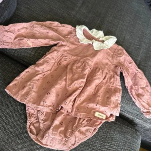 Rosa långärmad klänning från Newbie - Söt rosa klänning från Newbie med lång ärm och vit spetskrage. Klänningen har broderade detaljer och en volangliknande kjol. Tillverkad i mjuk bomull, perfekt för vår och sommar. Passar små barn och har en klassisk, gullig stil.