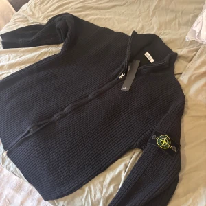 Svart stickad kofta från Stone Island - Svart grovstickad kofta från Stone Island med dragkedja framtill och ribbad krage. Klassisk logotyp-patch på vänster ärm. Tröjan har långärmad passform och är tillverkad i mjukt ullmaterial, perfekt för kyliga dagar. Storlek L passar m