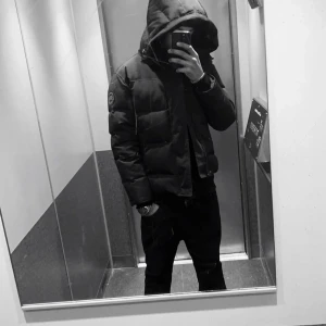 Svart pufferjacka från Canada Goose - Svart pufferjacka från Canada Goose med huva och broderad logotyp på ärmen. Jackan är quiltad och har en clean look, perfekt för kalla dagar. Modellen är windham M/M och har dragkedja framtill. Jackan är tillverkad i polyester och dun för extra värme.