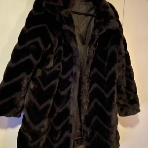 Svart pälsjacka med zigzag-mönster - Säljer en svart pälsjacka med snyggt zigzag-mönster. Jackan har en fluffig och lyxig känsla, perfekt för dig som vill sticka ut med en cool look. Den har lång ärm och är fodrad på insidan för extra komfort. Passar dig som gillar statement-plagg.