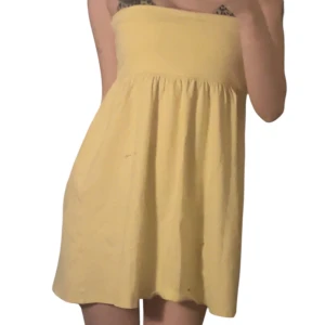 Gul axelbandslös kort klänning - Söt axelbandslös kort klänning i ljusgul färg med utsvängd kjol och markerad midja. Klänningen har en enkel och stilren design, perfekt för varma dagar. Tillverkad i mjukt bomullstyg som känns skönt mot huden.