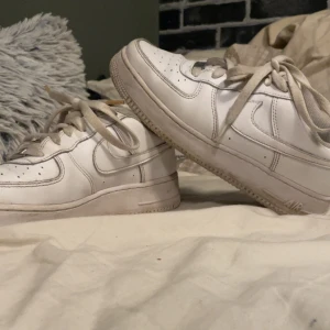 Nike Air Force 1 vita sneakers - Nike Air Force 1 sneakers i helvitt med klassisk design och perforerad tå. Skorna har snörning, platt sula och rund tå. Tillverkade i skinn med metall-detalj på snöret. Snygga och tidlösa, perfekta för dig som gillar streetstyle.