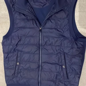 Mörkblå dunväst från Polo Ralph Lauren - Snygg mörkblå dunväst från Polo Ralph Lauren med quiltad framsida, två fickor med knappstängning och dragkedja framtill. Västen har en stilren design med slät baksida och diskret broderad logga på bröstet. Perfekt för lager-på-lager och chill stil.