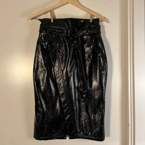  Pennkjol med bälte - Snygg svart midikjol i glansigt PU-material med hög midja och brett bälte med metallspänne. Kjolen har omlottkänsla och en liten slits framtill. Perfekt för dig som gillar statement-plagg och vill sticka ut med en edgy look.