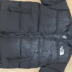 Svart pufferjacka från The North Face - Svart pufferjacka från The North Face i storlek M. Jackan har hög krage, två sidofickor med dragkedja och tydligt logotryck både fram och bak. Materialet är syntet och jackan har en quiltad design med dragkedja framtill.