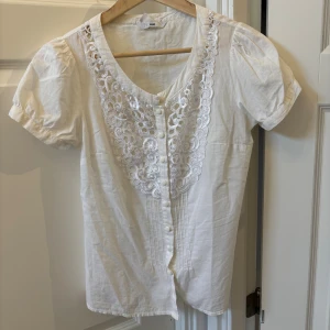 Vit blus med spets och knappar H&M - Söt vit blus från H&M med korta puffärmar, dekorativa knappar framtill och vacker spetsdetalj längs urringningen. OBS. Att den är genomskinlig! Skriv för fler bilder🥰