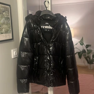 Svart glansig pufferjacka från Only - Svart pufferjacka från Only med glansig finish och huva. Jackan har breda, quiltade sektioner och dragkedja framtill. Perfekt för dig som vill ha en varm och snygg jacka med lite extra attityd. Passar dig som gillar streetstyle och vill sticka ut.