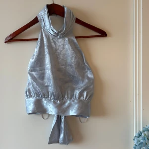 Ljusblå halterneck polotopp i satin - Snygg ljusblå polotopp i satin med blommigt mönster. Toppen har omlottknytning i ryggen, bred krage med knappar och en croppad passform. Materialet är glansigt och mjukt, perfekt för att sticka ut med en elegant vibe. Passar även strl M, aldrig använd 