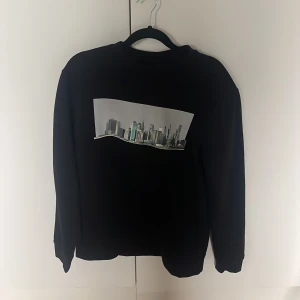 Svart sweatshirt  - Svart sweatshirt med tryck av en modern stadssilhuett på bröstet. Tröjan har rund hals och långa ärmar, perfekt för dig som gillar stilrena plagg med coolt print. Passar till jeans eller joggers för en avslappnad look.