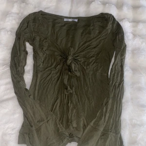 Olivgrön långärmad topp med knyt - Säljer en olivgrön långärmad topp från Doll House med snygg knytning framtill och v-ringad hals. Toppen har en lätt loose passform och är tillverkad i mjuk bomull. Perfekt för dig som gillar coola detaljer och jordnära färger.
