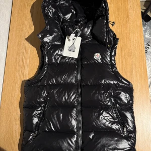 Svart dunväst från Moncler med huva - svart dunväst från Moncler med glansig finish, huva och dragkedja framtill. Västen har två fickor med dragkedja och klassisk Moncler-logga på bröstet. Nfc tagg fungerar.