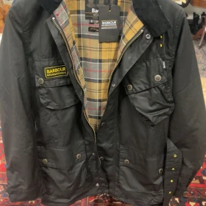 Svart Barbour International jacka S - Svart Barbour International jacka i slim fit med klassisk gul logotyp på bröstet. Jackan har flera fickor med tryckknappar, dragkedja och rutigt foder i grått, gult och rött. Perfekt för dig som gillar stilren och funktionell design.