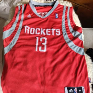 Houston Rockets Harden basketlinne Adidas - Röd Houston Rockets baskettröja med nummer 13 och namnet Harden på ryggen. Tröjan är ärmlös, har grå detaljer och broderade NBA- och Adidas-loggor. Tillverkad i ventilerande meshmaterial, perfekt för sport och streetwear.