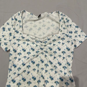 Vit blommig ribbad topp - Söt vit topp med blått blommönster och ribbad struktur. Toppen har korta ärmar, v-ringad halsringning med lätt rynk framtill och är figurnära i modellen. Perfekt för en fräsch och somrig look.