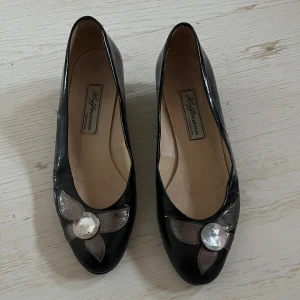 Svarta vintage pumps med dekoration - Snygga svarta pumps från Hoffmann med rund tå och låg klack. Skorna har en glansig finish och är dekorerade med en stor kristall och silvriga blad framtill. Perfekt för dig som gillar unika detaljer eller till nyår