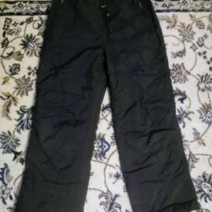 Svarta täckbyxor från Craft - Svarta täckbyxor från Craft med elastisk midja och snörning för skön passform. Byxorna har dragkedjeförsedda fickor och är gjorda i slitstarkt syntetmaterial, perfekta för kalla dagar och utomhusaktiviteter. Skandinavisk design med sportig vibe.