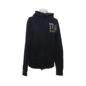 Svart hoodie från Hollister med päls - Svart hoodie från Hollister med vit och gul text på bröstet. Tröjan har dragkedja, huva med snörning och två fickor framtill. Tillverkad i bomull och polyester, perfekt för kyliga dagar.