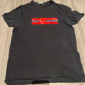 Svart Moncler t-shirt med röd logga - Svart t-shirt från Moncler i mjuk bomull med klassisk passform. Framtill finns en röd och blå randig logga med Moncler-tryck och på ärmen sitter det ikoniska broderade Moncler-märket. Perfekt för dig som gillar stilrena och exklusiva plagg.