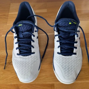 Vita och marinblå Asics sneakers - Snygga inneskor Asics sneakers i vitt och marinblått med gula detaljer på sulan. Skorna har snörning och är tillverkade i syntetmaterial och mesh för bra andningsförmåga. Perfekta för träning eller sportaktiviteter. Sulan är märkt med 'non marking'. Perfekt till inomhusidrott i skolan eller vid sport. Nästan oanvända
